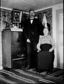 Ett äldre par i finrummets hörn vid en sekretär. Mannen står, och når nästan upp till det låga stugtaket, och kvinnan sitter. Rummets tapet är ovanlig med spridda blombuketter (blåklocka, primula, tusensköna) i naiv stil och en kurbitsliknande takbård. Mannen är finklädd med fluga och rova; hans högra hands fingrar ser ut att ha skadats.