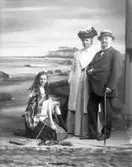 Familjebild, 1910. En familj bestående av en kvinna och en man samt en flicka. De vuxna är klädda i promenadkläder och mannen håller en käpp i handen, Flickan sitter på en sten. Hon har tagit av sig skorna och en av strumporna. Fonden föreställer en havsvik (Apelviken) och Varbergs fästning.