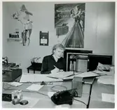 Kontoret på Monarks cykelverkstad. En kvinna sitter vid ett skrivbord och skriver i en pärm. På väggen bakom henne sitter en affisch med reklam för Monarks cyklar och en för Monarks radiomöbel 