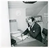 David Danielsson sitter och talar i telefon på Monarks kontor, klockan på väggen bakom honom visar på halv åtta. I bakgrunden står också en radio.