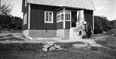 August och Tilda Snygg står med sina två döttrar på trappan till sitt hus, uppfört 1934. Grunden har synlig brädform och i vinkeln mot verandan med spröjsade fönster har en planerad rabatt kantats med tegelstenar ställda på högkant. Taket är belagt med stickspån (pärt).
