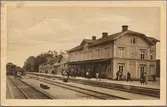Stationsområde med stationshus och resande på perrong.