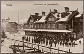 Stationsområde med resande på perrong och hotell i bakgrunden.