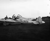 Ett litet propellerflygplan sett bakifrån står på ett gräsfält (Getterön?) och en man i overall förefaller tanka planet via vingen. En vagn med två slangförsedda behållare står bakom vingen.