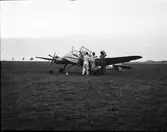 Ett litet propellerflygplan står på ett vidsträckt gräsfält och tre män i overaller arbetar vid dess ena vinge. En vagn med två slangförsedda behållare står intill dem och under den andra vingen står en box.