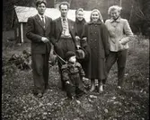 Ett sällskap står framför Persgårds stuga i skogen. Mannen längst till vänster heter Gösta Svahn och han håller lilla dottern Margareta i overall framför sig i en sele. Längst till höger står hans fru Inga och baktill står hennes syster Eva. Paret i mitten är Inga och Evas kusin Anna-Lisa och hennes make smeden Nils.