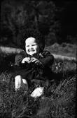 Ett litet barn sitter i gröngräset i mockasiner och manchesterdress med en stor veckad skärm på mössan. I händerna håller barnet ett runt metallföremål, kanske en klocka eller dosa av något slag.