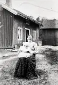 En äldre kvinna sitter utomhus på en stol hållandes ett litet barn i knäet, okänt årtal. I bakgrunden ses 