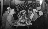 Glada middagsgäster under Arbogautställningen. Män och kvinnor som avslutat måltiden på en restaurang. De har dryck kvar i glasen. En stor blomsteruppsättning står på bordet.
Den fjärde mannen, i vänstra raden, kan möjligen vara Sven Lind.