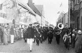 Livgrenadjärregementets musikkår från I3 i Örebro paraderar på Nygatan i samband med Riksdagens 500-årsjubileum i Arboga.
Publiken står efter husväggarna när den uniformerade blåsorkestern tågar förbi. Bland butiksskyltarna ses 