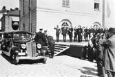 Kung Gustaf V och prins Wilhelm använder i bil till festligheterna i Arboga. Här ska firas Riksdagens 500-årsjubileum. Två män i uniform gör honnör. En scen är uppbyggd på Stora torget, intill Rådhuset.
(Arbogautställningen pågår samtidigt och kungen ska hinna besöka den också).