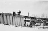 Här byggs den stora utställningshallen till Arbogautställningen. Tre män arbetar på taket och två män arbetar på en vägg. Invigningen ska ske 25 maj 1935. Nu är det vinter och snö.
I bakgrunden ses tornet på Heliga Trefaldighetskyrkan.