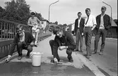 Arboga Tekniska Skola, skändning av nya elever. Per Bengtsson (till vänster) och en annan nykomling tvingas skura Herrgårdsbron med rotborste medan tre äldre elever övervakar arbetet. I bakgrunden ses tornet på Sankt Nikolai kyrka.