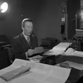 Arboga Tidning, interiör. En man, iklädd kavaj och slips, sitter vid ett skrivbord. Han har mycket papper omkring sig. Nu öppnar han ett kuvert. På bordet står en telefon. En annan telefon (möjligen en interntelefon) hänger på väggen.