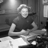 Arboga Tidning, interiör.
Bakom skrivbordet sitter Ann-Marie Eriksson med en penna i handen. Hon har två pappershögar framför sig. På bordet finns även en pärm, en telefon och en räknemaskin. Bakom hennes rygg, står en skrivmaskin, på en hylla.