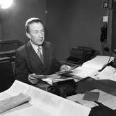 Arboga Tidning, interiör och personal.
En man, iklädd kavaj och slips, sitter vid ett skrivbord. Han har mycket papper på bordet. En telefon skymtar på skrivbordet. En annan telefon (kanske en interntelefon?) hänger på väggen. På ett bord, vid sidan, står en skrivmaskin.