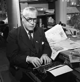 Arboga Tidning, personal och interiör. Redaktör Eitel Noll vid skrivmaskinen. Han har en uppslagen tidning och en telefon på skrivbordet. I bakgrunden syns en hylla med papper och föremål i. Han sitter nära fönstret, hans kontor ligger någon våning upp i byggnaden. Bakom hans rygg är dörren öppen in till nästa kontorsrum.