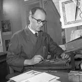 Arboga Tidning, personal och interiör.
En man arbetar med typer. Skjorta och slips syns under skyddsrocken. Han har ett pincettliknande verktyg i handen. På väggen, i förgrunden, syns en lång sax och en almanacka. På väggen bakom honom skymtar också en almanacka samt ett porträtt på kung Gustav Vl Adolf.