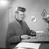 Arboga Tidning, personal och interiör.
Ung man, iklädd skyddsrock. Han är möjligen typograf. På väggen bakom honom sitter en bild på trumpetspelaren Louis Armstrong.