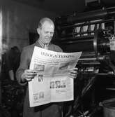 Arboga Tidning, personal och interiör.
En man håller i en färsk upplaga av tidningen. Han är klädd i arbetsrock och står intill en tryckpress. Två andra män skymtar i bakgrunden.