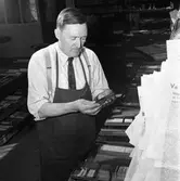 Arboga Tidning, personal och interiör.
Man, iklädd skjorta, slips och förkläde, arbetar omgiven av typer och ramar. Hans yrkestitel är förmodligen sättare.