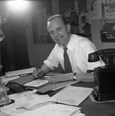 Arboga Tidning, interiör och personal.
En man sitter vid skrivbordet och skissar. Han har en penna i handen och ett papper framför sig. Han har många papper utspridda på bordet. Där finns även ett pennfat och en telefon. På väggen, bakom mannens rygg, ses flera affischer.
I fotots övre vänstra hörn, ses handen på den person som hjälper fotografen att hålla i blixten.