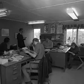 Arboga Tidning, personal och interiör. Det är full aktivitet på tidningsredaktionen; skrivmaskinerna smattrar vid tre skrivbord. Vid det fjärde sitter en person och läser igenom sina anteckningar. En man står upp och läser något på ett papper. Journalisterna har egna telefoner. Askkoppar och tobakspaket ses på skrivborden. Genom fönstren ser man att kontoret ligger på andra våningen. På väggen hänger affischer om det senaste från Riksteatern och biografen.