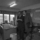 Arboga Tidning, personal och interiör. Tre personer vistas i rummet. Två personer står och tittar på ett papper som den ene mannen håller i. Mannen med papperet är klädd i kostym och slips. Den andre mannen, Willy Johansson, är klädd i överrock och har en portfölj under armen. Mannen vid skrivbordet röker pipa.
I rummet ses högar med tidningar och fack för papper.
På bordet, i förgrunden, står en kamera. En kaffekopp skymtar.