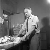 Arboga Tidning, personal och interiör. En man packar tidningar. Tidningsbuntar är travade i högar. Ytterdörren står öppen.