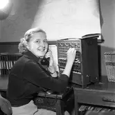 Arboga Tidning, personal och interiör. En kvinna sitter i telefonväxeln. Hon har en telefonlur i handen och kopplar samtalet med den andra. Till vänster om henne står en mängd pärmar. På väggen, framför henne, hänger en stadskarta.