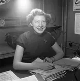 Arboga Tidning, personal och interiör. Vid skrivbordet sitter Ann-Marie Eriksson. Hon har en penna i handen och en bunt papper framför sig. Hon bär en brosch på jumpern. Bakom hennes rygg står en skrivmaskin.
I fotots övre, vänstra hörn syns handen på fotografens assistent, som håller i blixten.