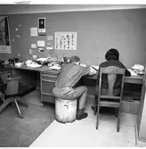 Arboga Tidning, personal och interiör. Två journalister arbetar vid samma skrivbord.  De har ryggen mot fotografen. Mannen sitter på en papperskorg och kvinnan på en trästol. Till vänster om dem finns en obemannad arbetsplats med en telefon och en arbetsstol på hjul. En skrivmaskin skymtar. På väggen sitter bland annat en affisch med olika svampar.