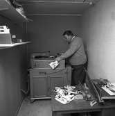 Arboga Tidning, personal och interiör. En man arbetar vid en maskin. I förgrunden syns ett bord med en skärmaskin, en borste och några bilder.