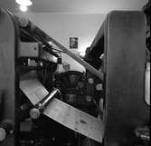Arboga Tidning, personal och interiör. Tryckeriet.
En man arbetar vid tryckpressen. Tryckta tidningssidor rullar mellan valsarna medan mannen kontrollerar maskinen.