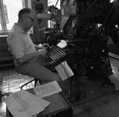 Arboga Tidning, personal och interiör. En man sitter vid en maskin. Maskinen har ett tangentbord som han skriver på. Vid sidan av mannen ligger papper utspridda.