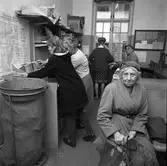 Arboga Tidning ska distribueras. Bilden kan möjligen vara tagen på järnvägsstationen eftersom TGOJ´s turlista sitter på väggen. Mannen med hatten har anknytning till Arboga Tidning. Kvinnorna, vid arbetsbordet, ägnar sig åt en tidningsbunt. Kvinnan vid fönstret har ryggen mot fotografen. Hon har en hög håruppsättning. I förgrunden sitter en kvinna på en bänk. Kanske väntar hon på tåget? Hon är iklädd mössa och en kappa med en svångrem om midjan. I handen håller hon en typisk matsäcksväska.