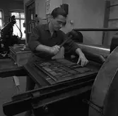 Arboga Tidning, personal och interiör. En man sätter en tidningssida. Han håller en gummiklubba i handen. På väggen hänger en almanacka. En stor maskin skymtar i bakgrunden.