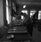 Arboga Tidning, personal och interiör. Två män som bygger tidningssidor med typer. Deras titel kan vara sättare. På väggen hänger en telefon. I förgrunden ses ett askfat. I bakgrunden står en stor maskin.