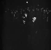 Arboga-natta, orientering. En glad man håller något i handen. Bakom ett band står publiken i ytterkläder. Bland publiken ses två män i uniform.