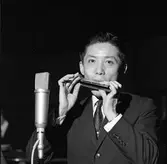 Arbogabor deltar, som publik, i radioprogrammet Frukostklubben, ett underhållningsprogram som sänds på lördagsmorgnar. En man, iklädd kostym och slips, spelar munspel. En mikrofon står intill.