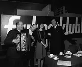 Arbogabor gästar radioprogrammet Frukostklubben, ett underhållningsprogram som sänds på lördagsmorgnar. En man undersöker Andrew Walters dragspel. En glad kvinna står intill Sigge Fürst, den ständige programledaren.