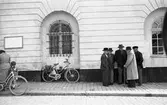 Fem äldre män har träffats på gatan. Fyra bär hatt och en bär keps. En man har käpp. 
Byggnaden inhyser polisstationen och ett av fönstren är försett med järngaller. Under fönstret står en cykel med packväskor. Till vänster i bild skymtar en kvinna, ett barn och en cykel.