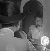 Arbogarevyns medlemmar spelar upp sin Jubileumsrevy. Rune Lindqvist sitter i logen och sminkar sig framför spegeln.
Han är iklädd stormhatt och fluga.