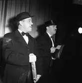 Medlemmarna i Arbogarevyn spelar upp sin Jubileumsrevy. Två herrar på scenen, den högra är Rune Ström. De är klädda i kostym, fluga och hatt. Rune har en portfölj under armen.