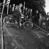 Arbogas Stjärnknutte
Tävling i terrängkörning med motorcykel. Här står motorcykeln på tvären i nerförsbacken. Publiken tittar intresserat på.
