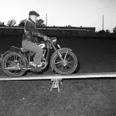 Arbogas Stjärnknutte
Balansövning med motorcykel, körning på lutande bräda. Tävling.
Mannen är iklädd keps och skinnjacka.