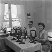 Börjesson tillverkar Arbogaspegel
Ett par sitter vid köksbordet. Flera spegelramar står uppställda framför dem. Kvinnan har förkläde på sig. På väggen hänger en almanacka. Genom fönstret syns villor.