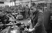 Arne Engström,iklädd skyddsrock, vid en maskin. Miljön är fabriksinteriör. I bakgrunden ses två kvinnor.