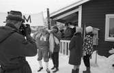 Skidtävlingen AT-loppet.
En man i krimmelmössa håller kläder och en termos i famnen. I förgrunden står en ung man med mössa. Ett par stavar syns i bakgrunden.
(AT kan betyda Arboga Tidning)