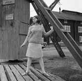 Ateljé Touche. En ung kvinna visar mode; jumper och kjol.
Bilden är tagen i hamnen med hamnmagasin i bakgrunden.